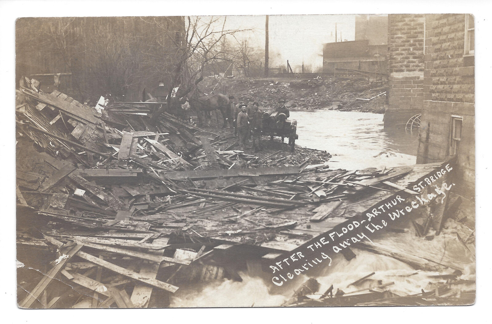 Historical postcard showing people, horses, a cart and debris beside Arthur Street Bridge after a flood.